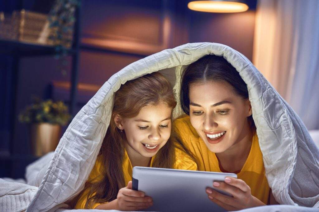 mom and daughter under the duvet cover using a tablet that is emitting blue light